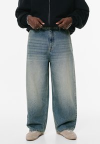 Svijetloplavi traperice širokih nogavica, s visokim strukom, izblijedjelom teksturom i suptilnim naborima blizu prednjih džepova.