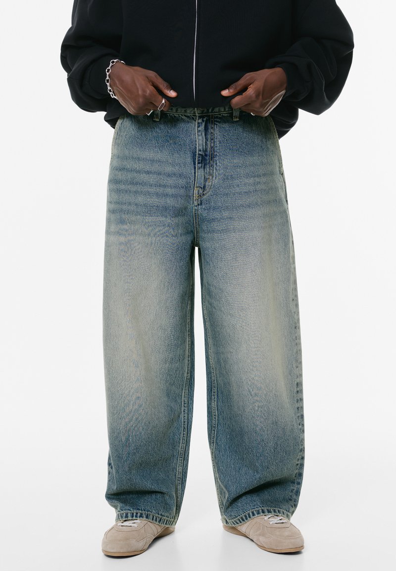 Svijetloplave, široke traperice od denima, s visokim strukom, izblijedjelom teksturom i suptilnim sitnim naborima blizu prednjih džepova.