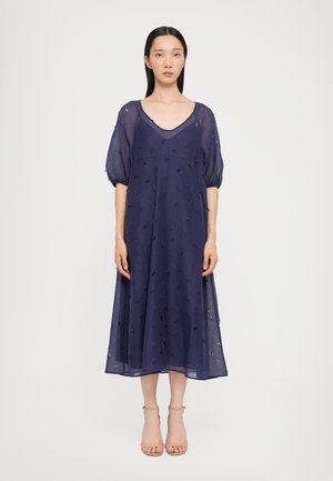 Marineblauwe jurk tot de knie met doorzichtige stof, V-hals, opgezette mouwen en geborduurde bloemdetails overal. Gedragen met nude hakken.