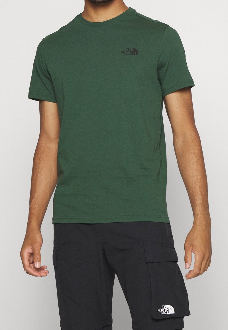 Grön t-shirt i bomull med korta ärmar, rundad halsringning och en liten svart logotypdesign på vänster bröst. Bärs med svarta cargo-shorts.