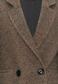 Ruskea ja musta herringbone-kuvioinen bleiseri, jossa on leveät läpät ja kaksi suurta mustaa nappia mustan sisävaatteen päällä.