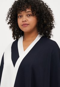 Námořnický top ve stylu kimona s bílými akcenty, s hlubokým V-výstřihem a hladce splývajícím materiálem. Krepaté vlasy rámující obličej, neutrální pozadí.
