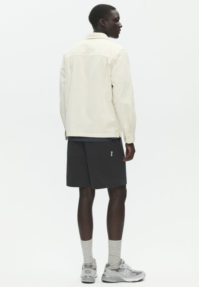 Homme vu de dos portant une veste crème clair, un short noir, des chaussettes grises et des baskets New Balance grises sur un fond blanc.