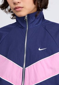 Mornarska vetrovka z visokim ovratnikom, rožnatim V-jem, belim logotipom Nike in srebrnim zadrgo. Gladka, lahka tkanina s športnim slogom.
