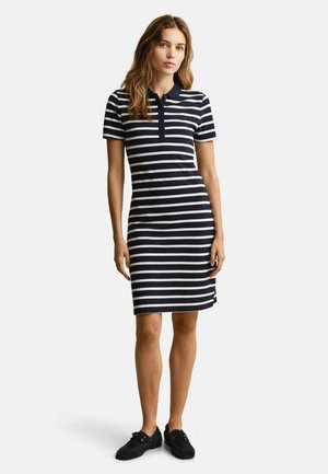 Vrouw staat, draagt een knielange polojurk met horizontale marineblauwe en witte strepen, korte mouwen en zwarte casual schoenen.