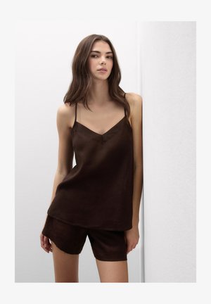 Young woman with long brown hair wearing a dark brown sleeveless top and matching shorts, standing against a white wall.
