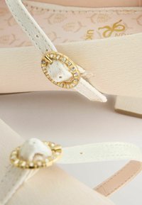 Pantofi bej cu talpă plată și finisaj texturat. Prezintă curele decorate cu accesorii circulare aurii și strasuri transparente.