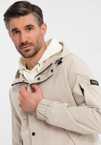 Mann mit kurzem, dunklem Haar und Bart, der eine beige Jacke über einem weißen Hoodie trägt, den Kragen der Jacke haltend und in die Kamera blickend, vor einem einfarbigen Hintergrund.