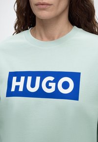 Femme portant un sweat-shirt vert clair avec un logo rectangulaire bleu indiquant "HUGO" en lettres blanches sur la poitrine.