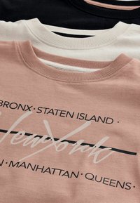Tre tröjor har en rundad halsringning. Färgerna inkluderar svart, kräm och rosé, med tryckt text och grafiska element på framsidan.