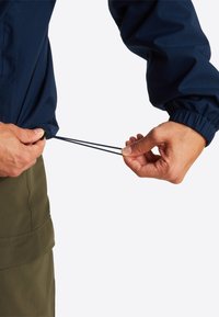 Coupe-vent bleu marine avec poignets élastiques ; une personne ajuste le cordon de serrage à l'ourlet, mettant en valeur un tissu lisse et léger ainsi qu'un design fonctionnel.