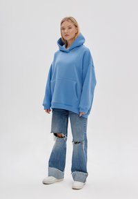 Felpa oversize blu chiaro realizzata in misto cotone, con tasca anteriore e polsini a costine. Abbinata a jeans a zampa d'elefante blu strappati e sneaker bianche.
