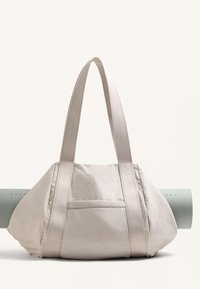 Ljusbeige väska med en mjuk textur, utrustad med två breda remmar och en sidoficka. En yogamatta i ljusgrön ligger lutad mot den.
