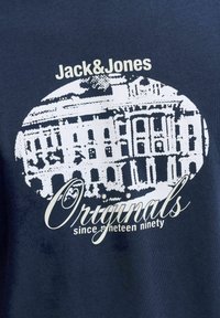 Tejido azul oscuro con un gráfico blanco de un edificio, texto "Jack&Jones" arriba y "Originales desde mil novecientos noventa" debajo del gráfico.