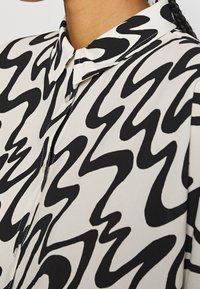 Černobílá halenka s vzorem, límečkem, hladkou látkou a zapínáním na knoflíky, s vlnitým abstraktním designem po celé ploše.