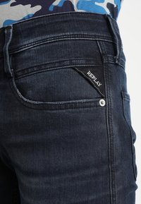 Mörkblå jeans med slim fit, med en distinkt vävd etikett märkt "REPLAY" på bakfickan och detaljer i silverfärg.