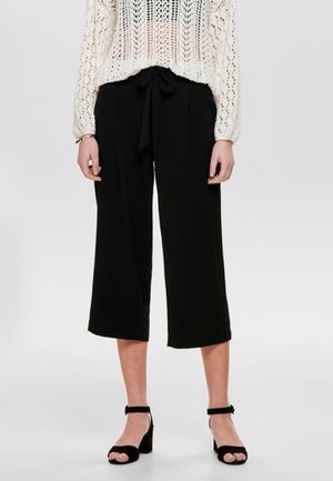 Mujer vistiendo pantalones anchos negros de pierna cropped con cintura anudada y sandalias negras de tacón bloque y punta abierta, combinados con un suéter blanco de punto.