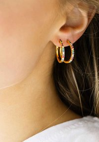 Orecchini a cerchio dorati decorati con strass multicolori, grandi e circolari, indossati su un orecchio di un modello con un tono di pelle uniforme.