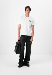 White cotton t-shirt with a navy blue floral logo on the chest, paired with black wide-leg jeans and a black tote bag.
