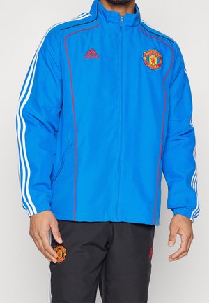 Mężczyzna noszący niebieską kurtkę Adidas z logo Manchester United i czarne spodnie z emblematem Manchester United na udzie.