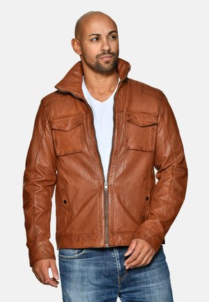 Braune Lederjacke mit stehendem Kragen, Reißverschluss und zwei Brusttaschen. Weiche Textur und taillierte Passform, kombiniert mit einem weißen Hemd.