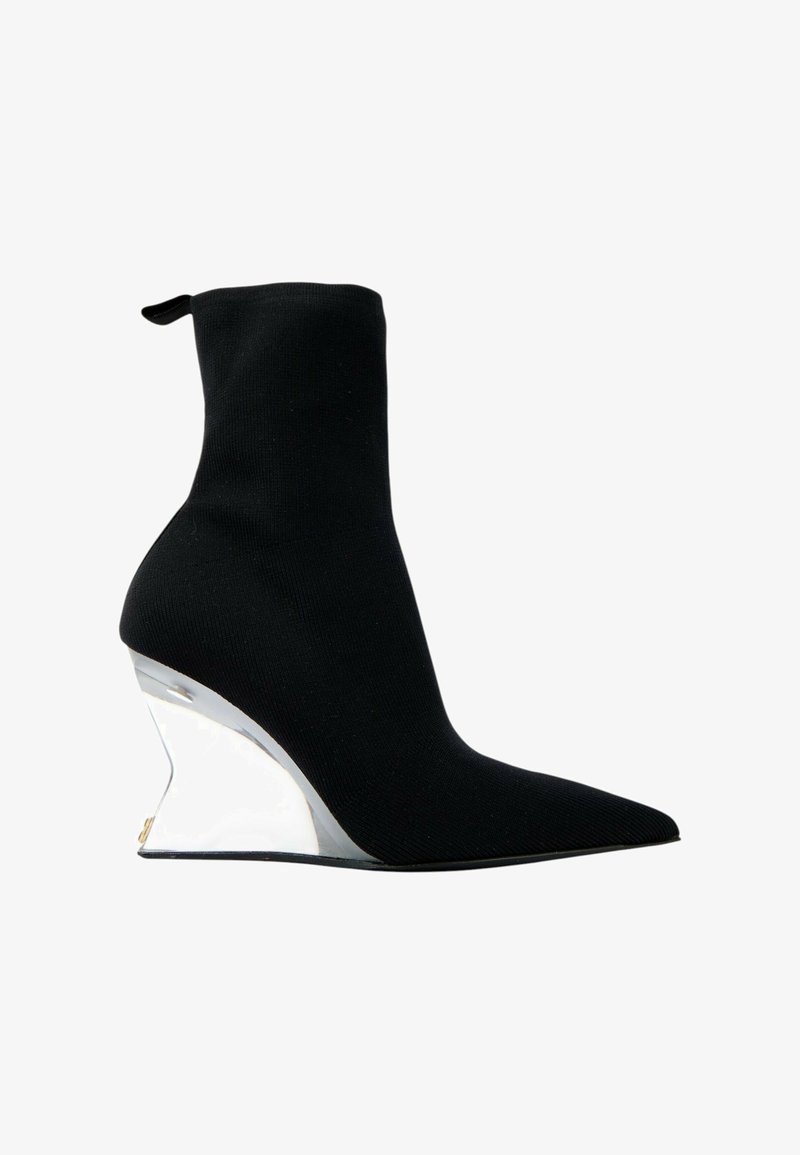 Bottine chaussette noire à bout pointu et talon compensé incurvé argenté réfléchissant sur fond blanc.