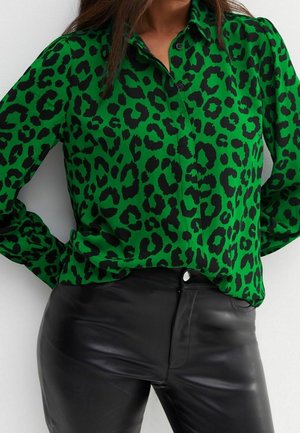 Kobieta w zielonej, długorękawowej koszuli zapinanej na guziki z czarnym wzorem w panterkę, zestawionej z czarnymi, skórzanymi spodniami z wysokim stanem.