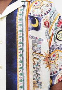 Πουκάμισο με πολύχρωμα, λεπτομερή μοτίβα που περιλαμβάνουν ημισέληνο, ήλιο, γεωμετρικά σχήματα και τη λέξη «MISBHV» με διακοσμητικά γράμματα.