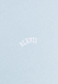 Helles blaues Stoffmaterial mit glatter Textur, das nahe der Mitte weißen, gestickten Text "BLKVIS" in einem gewölbten Stil zeigt.