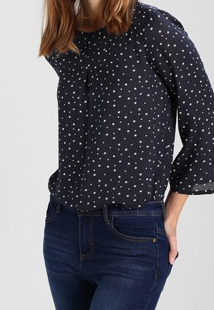 Kvinde iført en marineblå bluse med hvide stjernemønstre og trekvartærmer parret med mørkeblå jeans, hånd hvilende i baglommen.