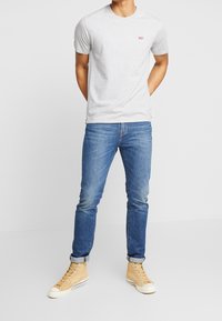 Svetle šedé bavlněné tričko s malým červeným logem, v kombinaci s modrými džíny a béžovými vysokými teniskami. Jednoduchý, ležérní střih.