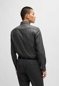 Chemise grise à boutons avec un motif texturé, manches longues et col pointu, associée à un pantalon gris ajusté et une ceinture noire.