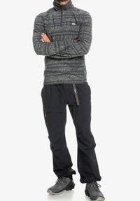 Polarowy sweter w ciemnoszarym kolorze z geometrycznymi wzorami; czarne spodnie z bocznymi kieszeniami. Posiada zamki błyskawiczne i regulowany pas.