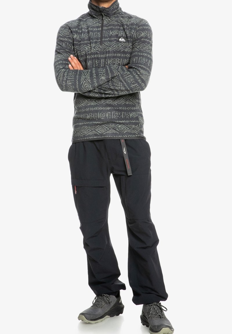 Polarowy sweter w ciemnoszarym kolorze z geometrycznymi wzorami; czarne spodnie z bocznymi kieszeniami. Posiada zamki błyskawiczne i regulowany pas.