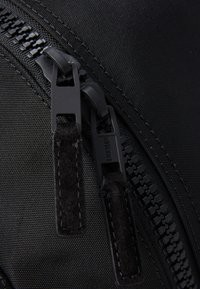 Sort nylonskæring med to trækstænger, der har struktureret stof og kontraststing. Lynlåsen har branding detaljeret på skyderen.