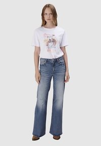 Weißes Grafik-T-Shirt mit Cowboy-Motiv, kombiniert mit hellblauen, hochtaillierten Schlagjeans mit Used-Details. Braune Stiefel.