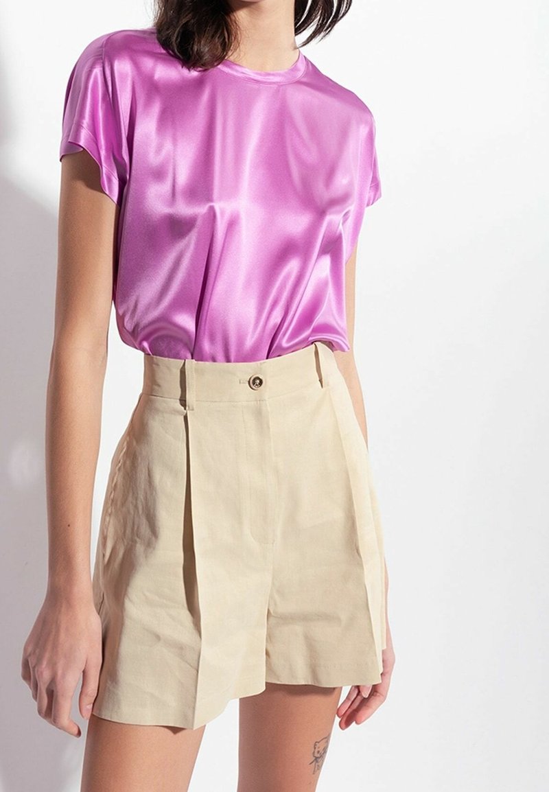 Femme portant un haut rose brillant à manches courtes rentré dans un short beige plissé taille haute, sur un fond clair uni.