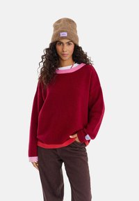 Czerwony sweter dziany z różowymi i koralowymi akcentami w formie prążków, zestawiony z brązowymi spodniami. Brązowa czapka typu beanie z fioletową metką. Miękka faktura.