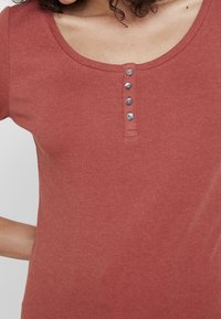 Frau in einem rostfarbenen Kurzarmhemd mit Rundhalsausschnitt und vier kleinen Knöpfen an der Vorderseite.