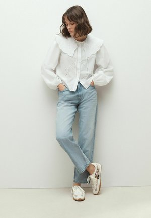 Frau mit braunen Haaren, die eine weiße Bluse mit großem besticktem Kragen, hellblaue, hoch taillierte Jeans und weiße Sneakers mit Leopardmuster trägt.