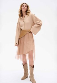Beżowy sweter z wyraźnymi guzikami i szerokimi rękawami, noszony z brązowym skórzanym paskiem na przezroczystej, warstwowej spódnicy z tiulu do kolan. Brązowe kowbojki.