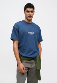 Μπλε βαμβακερό T-shirt με κοντά μανίκια και στρογγυλή λαιμόκοψη, με το λογότυπο "BARBOUR INTERNATIONAL" σε λευκό. Συνδυάζεται με πράσινο πουλόβερ δεμένο στη μέση.