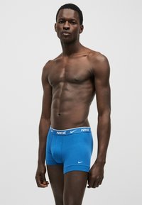 Αθλητικός μαύρος άνδρας όρθιος, φοράει μπλε μποξεράκια Nike με λογότυπο και λάστιχο στη μέση σε λευκό χρώμα, απλό ανοιχτό γκρι φόντο.