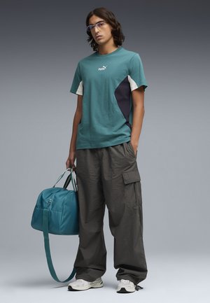 Jeune homme portant des lunettes, un T-shirt Puma bleu sarcelle et un pantalon cargo sombre ample, tenant un sac de sport bleu sarcelle, debout sur un fond gris.