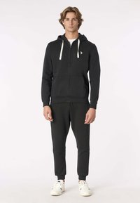 Felpa con cappuccio nera con zip e coulisse bianche, abbinata a pantaloni jogger neri. Entrambi in tessuto liscio con polsini e vita a coste, indossati con sneakers bianche.