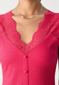 Femme portant un haut côtelé rose vif avec des détails en dentelle autour du col en V et deux boutons recouverts de tissu à l'avant.