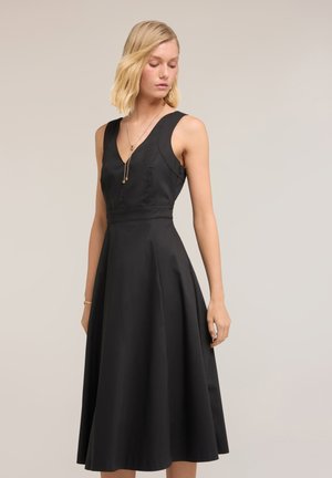 Femme blonde portant une robe midi noire sans manches avec un col en V, debout et regardant vers le bas sur un fond clair uni.