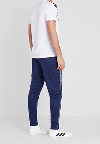 Man som står upp, iklädd marinblå träningsbyxor med vita sidostriper, vita sneakers med svarta striper och en vit kortärmad skjorta.