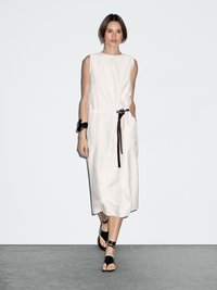 Woman wearing sleeveless white midi dress with black belt tie, black bracelet, and black ankle-wrap sandals, standing against plain white wall.