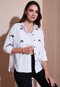 Mujer con cabello oscuro lleva una camisa blanca abotonada con patrones negros en forma de X y pantalones negros, posando contra una pared beige texturizada.